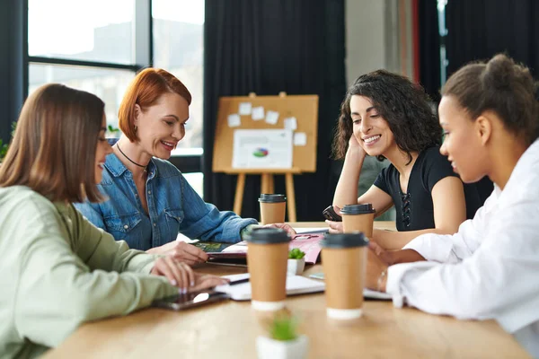 Cheerful redhead woman reading magazine to smiling multiethnic female friends while spending positive time with takeaway drinks in women interest club, communication and knowledge-sharing concept — Stock Photo