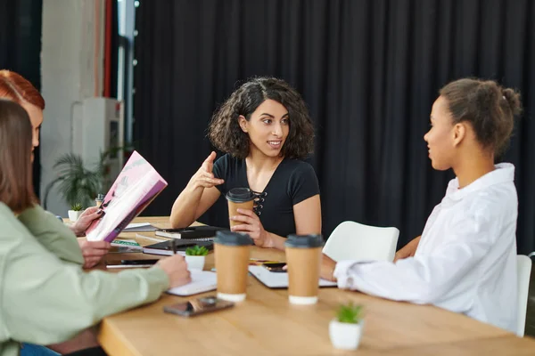 Smiling multiethnic woman holding coffee to go and talking to african american friend near members of interest club reading magazine in cozy atmosphere, communication and knowledge-sharing concept — Stock Photo