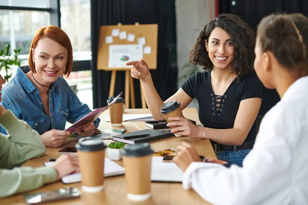 Allegra donna multirazziale che tiene il caffè per andare e punta con il dito mentre parla con amico afroamericano vicino ai membri del club di interesse, tempo libero e comunicazione in un'atmosfera accogliente — Foto stock