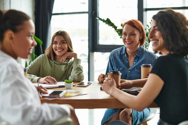 Mulheres felizes olhando para alegres namoradas multiétnicas sentadas perto de café para ir e falar no clube de interesse em primeiro plano turvo, lazer e comunicação em ambiente acolhedor — Fotografia de Stock