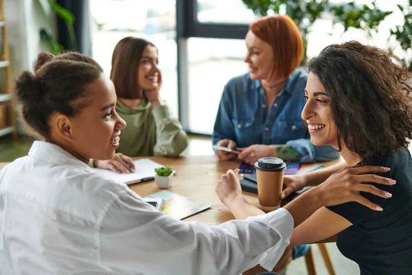 Cheerful multiracial woman with takeaway drink looking at african american girlfriend with braces near women talking on blurred background in interest club, communication in cozy atmosphere — Stock Photo