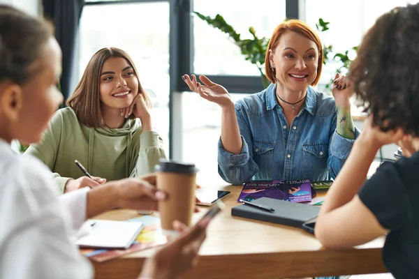 Excited redhead and tattooed woman gesturing and talking to multiethnic girlfriends near magazines on table in women interest club, blurred foreground, knowledge-sharing and socializing concept — Stock Photo