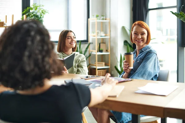 Optimistic women sitting with notebook and takeaway drink and smiling near female friend on blurred foreground during communication in interest club, knowledge-sharing and socializing concept — Stock Photo
