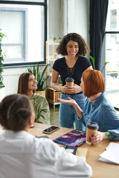 Redhead woman pointing at smiling multiracial friend standing with coffee to go near multicultural members in cozy atmosphere of interest club, spending time in friendly female community — Stock Photo