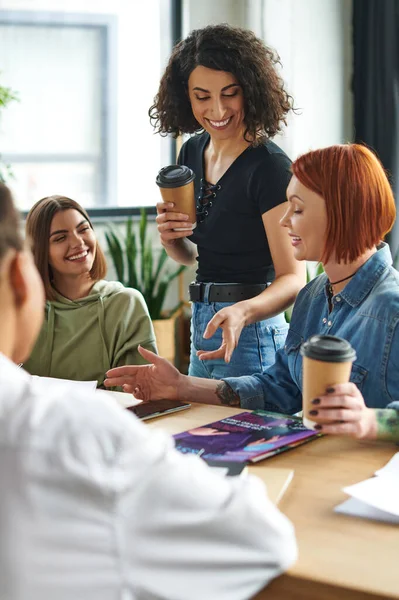 Joyful multiracial woman holding coffee to go and pointing at magazines near cheerful female friends talking in women interest club, spending time in friendly diverse community — Stock Photo