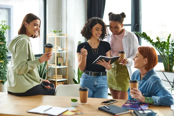 Happy multiethnic woman showing diary to african american girlfriend next to members of women club drinking takeaway coffee at table with magazines and notebooks, friendly diverse community — Stock Photo