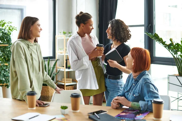 Pleased african american and multiracial women with notebook and paper cup talking near friends while spending time in interest club, happy gathering and leisure of diverse female team — Stock Photo