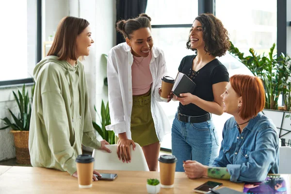 Overjoyed multiracial and african american girlfriends with notebook and takeaway drink laughing near positive women in interest club, happy gathering and leisure of diverse female team — Stock Photo