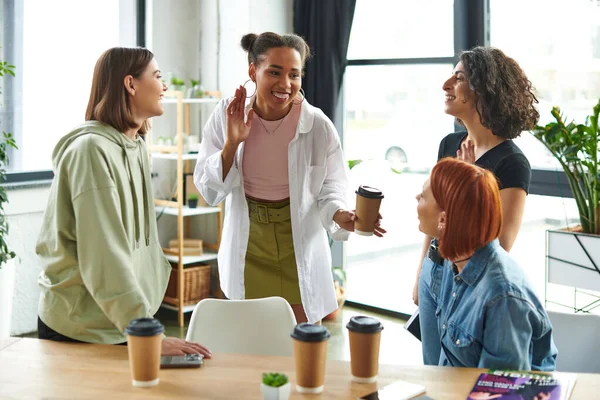 Joyful african american woman with takeaway drink showing new hoop earrings to multiethnic girlfriends in cozy atmosphere of women interest club, happy gathering and leisure of diverse female team — Stock Photo