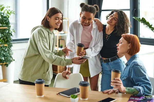 Joyous young woman showing mobile phone to multicultural friends near takeaway drinks and magazines on table in interest club, happy gathering and leisure of diverse female team — Stock Photo