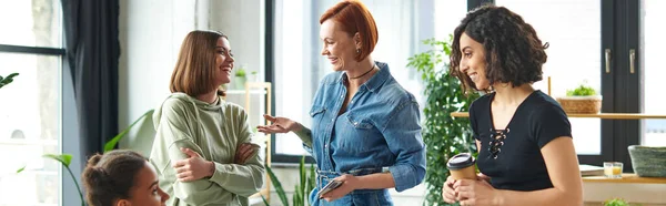 Redhead woman with coffee to go pointing with hand while talking to carefree friend near multiethnic members of interest club, happy gathering and leisure of diverse female team, banner — Stock Photo