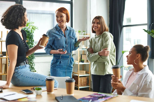 Overjoyed redhead woman standing with mobile phone, gesturing and talking to multiethnic friends holding takeaway drinks while spending time in interest club, leisure of diverse female team — Stock Photo