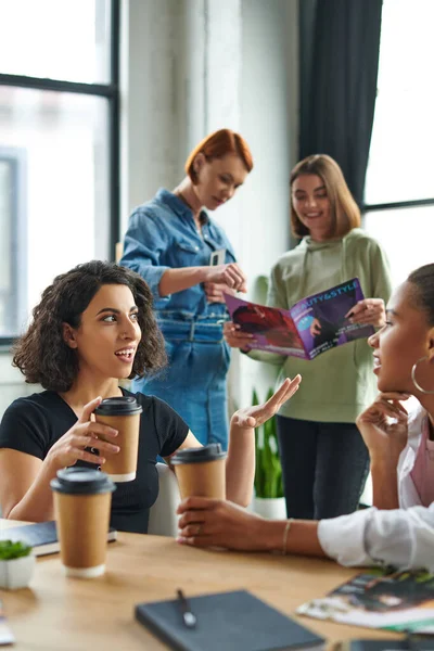 Multiracial woman with takeaway drink gesturing and talking to african american girlfriend near members of interest club reading magazine on blurred background, solidarity and understanding concept — Stock Photo
