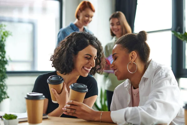 Overjoyed african american and multiracial female friends holding coffee to go and laughing with closed eyes near women on blurred background in interest club, solidarity and understanding concept — Stock Photo