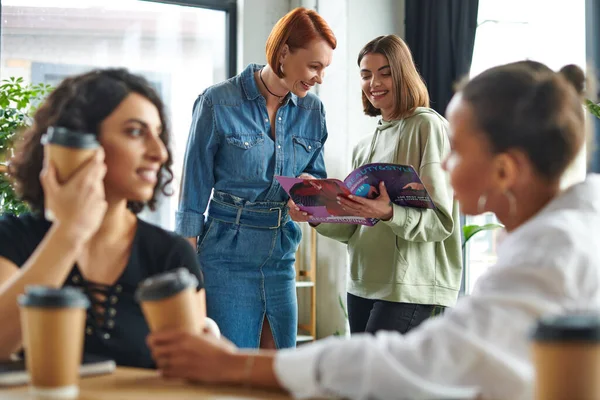 Cheerful girlfriends reading magazine while multiethnic women drinking coffee from paper cups and talking on blurred foreground in interest club, solidarity and understanding concept — Stock Photo