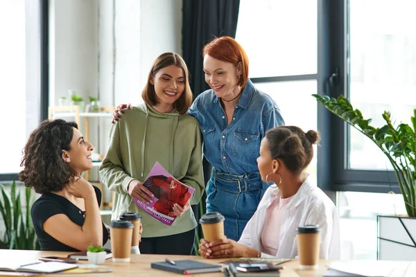 Optimistic redhead woman hugging young girlfriend standing with magazine near multicultural women and paper cup with takeaway drinks on table in interest club, solidarity and understanding concept — Stock Photo