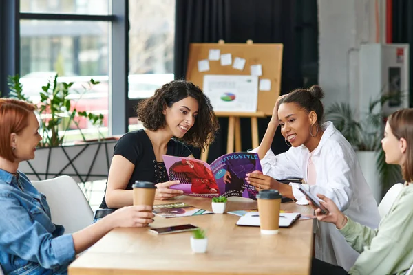 Happy african american and multiracial women reading beauty and style magazine near girlfriends sitting with smartphone and coffee to go in women club, common interests and knowledge-sharing concept — Stock Photo