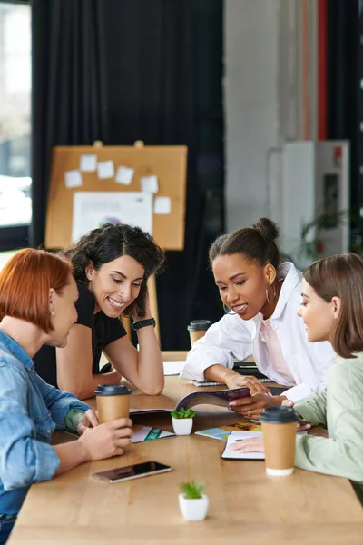 Des amies multiethniques inspirées passent du temps dans un club de femmes et un magazine de lecture tout en s'asseyant près de tasses en papier avec des boissons à emporter, des intérêts communs et un concept de partage des connaissances — Photo de stock