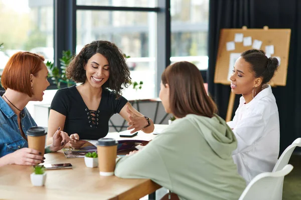 Excited multiracial woman gesturing while reading magazine to multicultural girlfriends sitting at table with coffee to go in women club, common interests and knowledge-sharing concept — Stock Photo