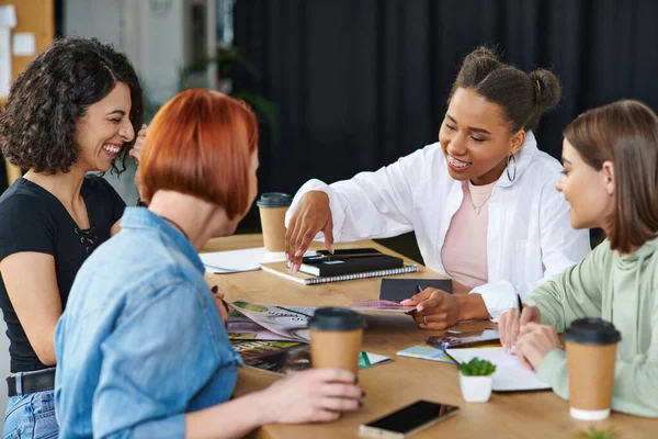 Joyful african american woman pointing at magazines while spending happy time with multicultural girlfriends near coffee to go in women club, common interests and knowledge-sharing concept — Stock Photo