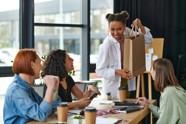 Young, smiling african american woman showing shopping bag to diverse group of multiethnic female friends sitting near paper cups and notebooks in interest club, sharing joy and positive emotions — Stock Photo