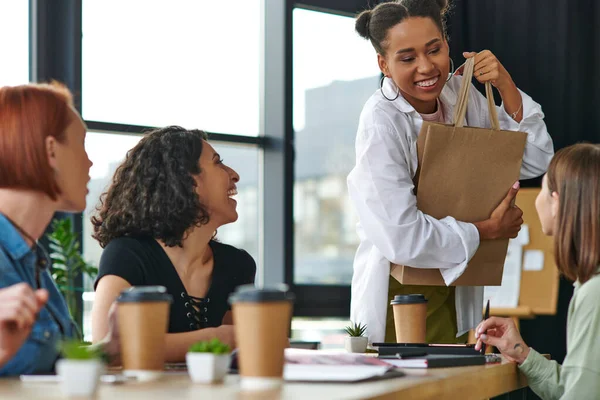 Young and overjoyed african american woman holding shopping bag near laughing multiethnic female friends sitting in women club at table with takeaway drinks, sharing joy and positive emotions — Stock Photo