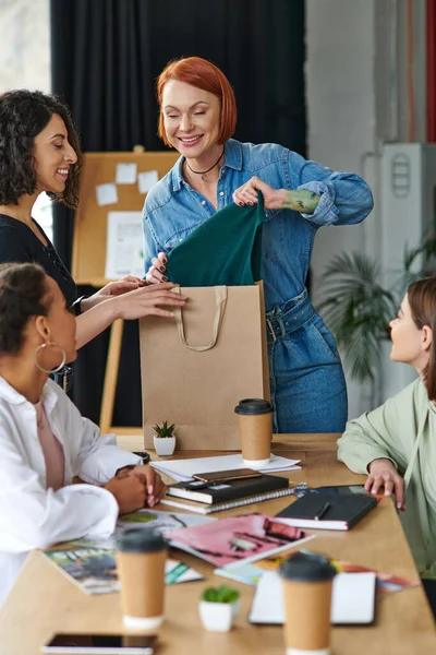 Felice rossa donna mostrando nuovo acquisto alle amiche multiculturali trascorrendo del tempo in un'atmosfera accogliente del club di interesse delle donne, condividendo gioia ed emozioni positive — Foto stock