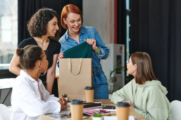 Redhead woman with shopping bag showing purchase to smiling multiethnic girlfriends near paper cups with takeaway drinks in cozy atmosphere of women interest club, sharing joy and positive emotions — Stock Photo