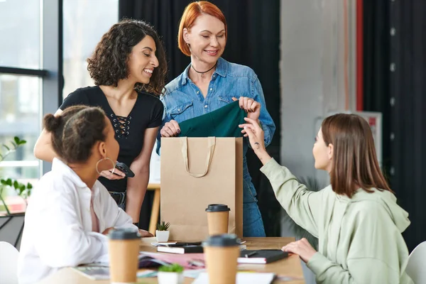 Pleased redhead woman showing new purchase to multicultural members of women interest club near paper cups with coffee to go on blurred foreground, sharing joy and positive emotions — Stock Photo