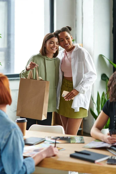 Joyous multiethnic girlfriends standing with mobile phone and shopping bag near members of women interest club sitting on blurred foreground, sharing joy and positive emotions — Stock Photo