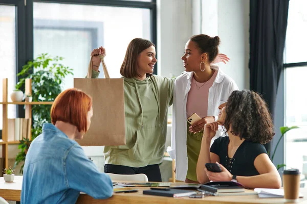 Excited woman showing shopping bag and smiling at african american girlfriend near multiethnic women sitting at table in friendly atmosphere of interest club, sharing joy and positive emotions — Stock Photo
