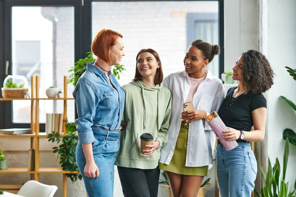 Satisfied multiethnic girlfriends in stylish casual clothes, with coffee to go, magazine and smartphone smiling at each other in women interest club, mutual support, solidarity and trust concept — Stock Photo