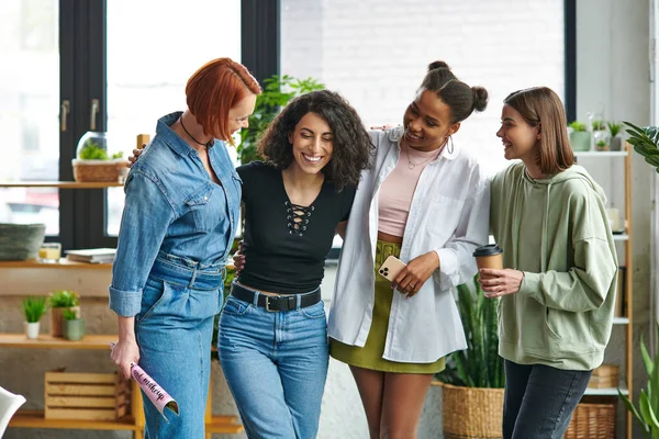 Overjoyed and stylish multicultural women with magazine, mobile phone and takeaway drink embracing in trendy atmosphere of women interest club, mutual support, solidarity and trust concept — Stock Photo