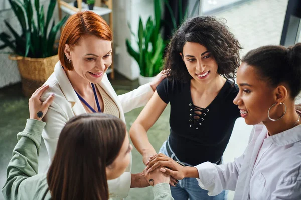 High angle view of positive redhead psychologist and diverse group of multiethnic women joining hands during motivation coaching, moral support and mental wellness concept — Stock Photo