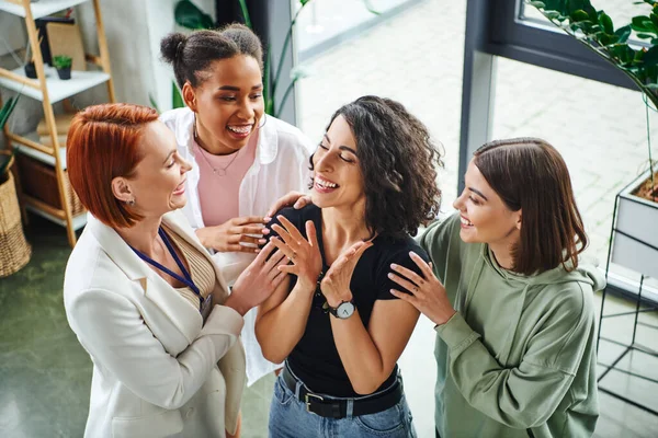 High angle view of happy motivation coach and multicultural girlfriends embracing happy multiracial woman smiling and gesturing with closed eyes, moral support and mental wellness concept — Stock Photo