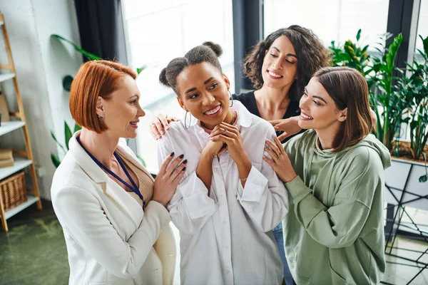 Positive psychologist and multiracial female friends hugging pleased african american woman during psychological coaching, moral support and mental wellness concept — Stock Photo