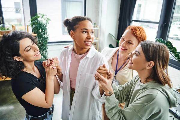 Multiethnic friends and smiling motivation coach holding hands of cheerful african american woman during supportive therapy in consulting room, moral support and mental wellness concept — Stock Photo
