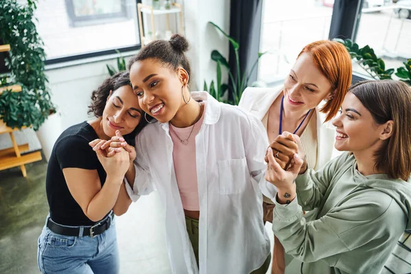 Smiling multiethnic female friends and motivation coach holding hands of happy african american woman while standing in psychology consulting room, moral support and mental wellness concept — Stock Photo