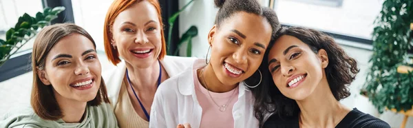 Joyful multicultural female friends looking at camera near motivation couch during psychology session in consulting room, moral support and mental wellness concept, banner — Stock Photo