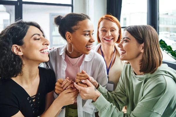 Mulheres multiétnicas alegres de mãos dadas de namorada afro-americana rindo com aparelhos perto de psicólogo sorridente em consultório, apoio moral e conceito de bem-estar mental — Fotografia de Stock