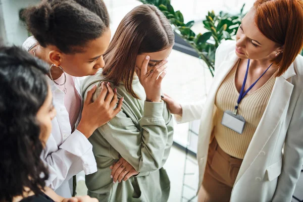 Young and depressed woman obscuring face with hand near multiethnic female friends and psychologist in consulting room, problem-solving and mutual support concept — Stock Photo