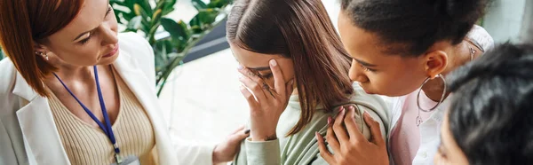 Young depressed woman obscuring face and crying near motivation coach and multiethnic friends during psychology session in consulting room, problem-solving and mutual support concept, banner — Stock Photo