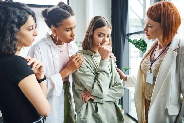 Redhead psychologist and multiethnic female friends looking at upset and thoughtful woman standing in consulting room, problem-solving and mutual support concept — Stock Photo