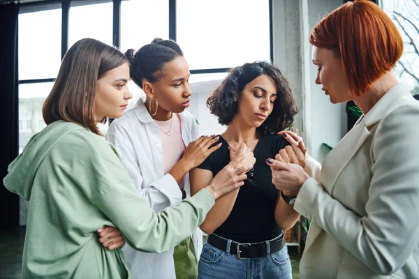 Upset multiracial woman standing with closed eyes near multiethnic friends and motivation coach supporting her during psychology session, problem-solving and psychological help concept — Stock Photo