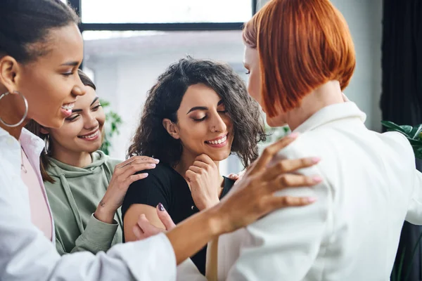 Happy multiracial woman smiling with closed eyes near positive multicultural girlfriends and redhead motivation coach in consulting room, self-improvement and mental health concept — Stock Photo