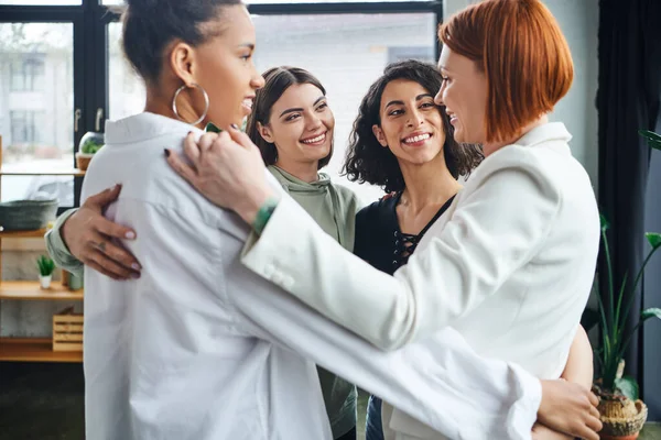 Happy and optimistic multicultural girlfriends embracing with redhead motivation coach and smiling at each other during supportive therapy in consulting room, female unity and support concept — Stock Photo