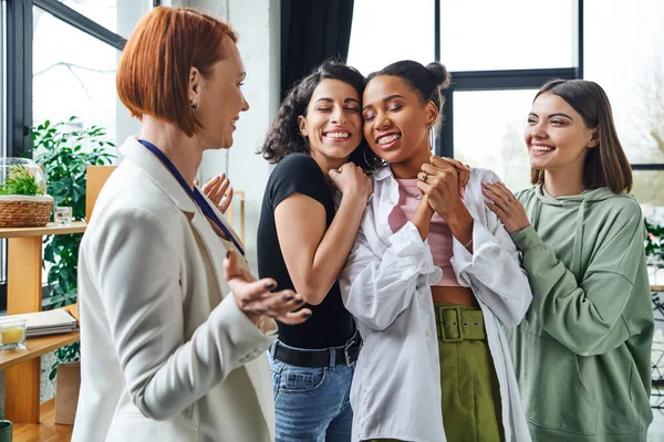 Cheerful multiethnic women embracing african american girlfriend near happy motivation coach gesturing in consulting room, female unity and support concept — Stock Photo