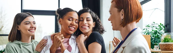 Excited multicultural women hugging multiracial girlfriend near pleased motivation coach during psychology session in consulting room, female unity and support concept, banner — Stock Photo