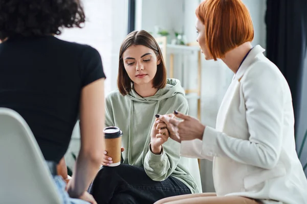 Redhead psychologist holding hand of young and sad woman sitting with coffee to go during supportive therapy in consulting room, problem-solving and mental wellness concept — Stock Photo