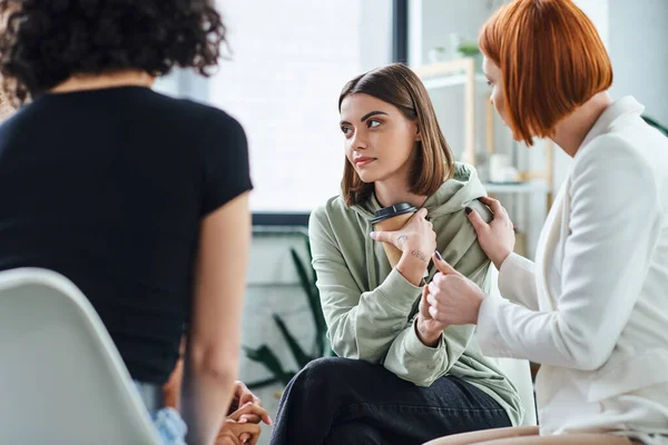 Redhead psychologist calming young upset woman sitting in consulting room with takeaway drink near girlfriend on blurred foreground, understanding and mutual support concept — Stock Photo